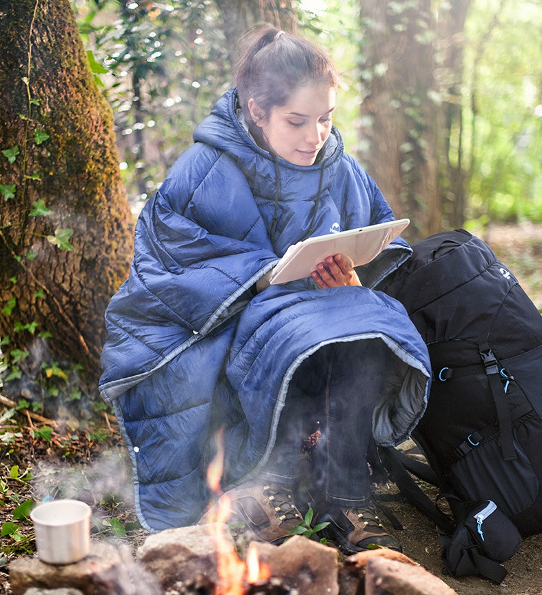 Person wearing blue wearable travel blanket cloak sitting by campfire in forest.